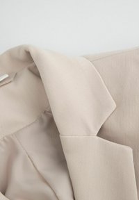 Beżowa marynarka z teksturowanej tkaniny, z klasycznym kołnierzem i widocznymi szwami. Wnętrze jest wyłożone gładkim materiałem.