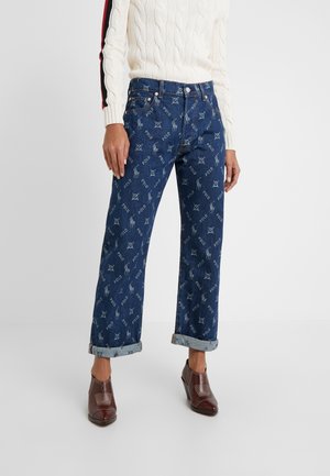 Persona con jeans azul oscuro con dobladillo y patrones del logo de Polo, y zapatos marrones con tacón, combinados con un suéter blanco de punto trenzado.