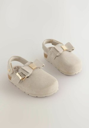 Sabots beige clair avec un nœud et des accents dorés. Ils présentent une sangle au dos et une semelle en liège, positionnés sur un fond clair.