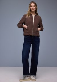 Kobieta stojąca z rękami w kieszeniach, ubrana w brązową kurtkę na zamek, ciemnoniebieskie szerokie dżinsy i białe sneakersy na szarym tle.