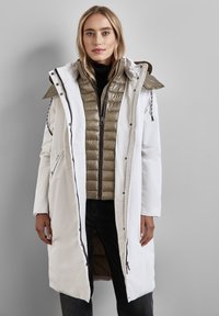 Lång vit ytterjacka med huva, lager över en vadderad olivgrön innerjacka. Har dragkedjor, fickor och kontrasterande sömmar.