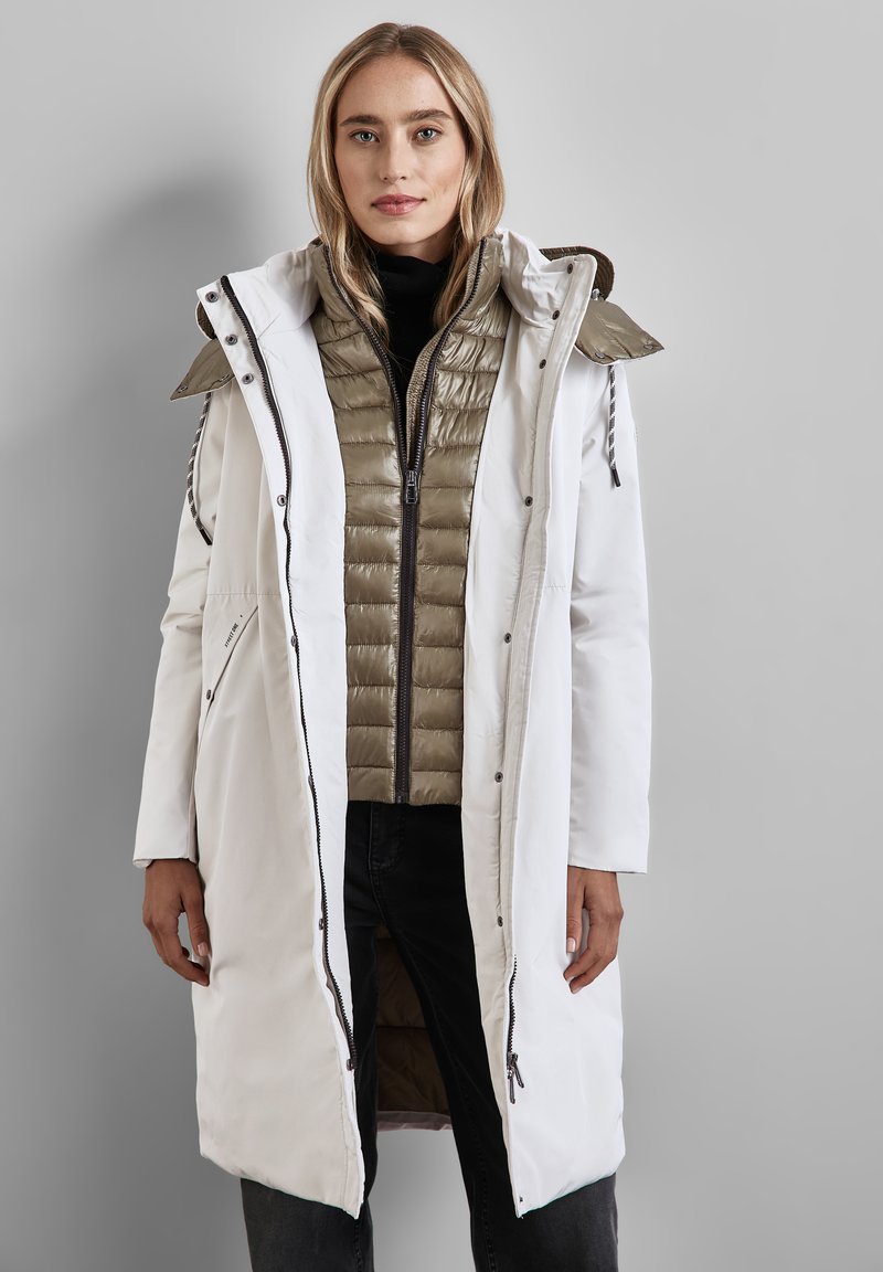Lång vit ytterjacka med huva, lager över en vadderad olivgrön innerjacka. Har dragkedjor, fickor och kontrasterande sömmar.