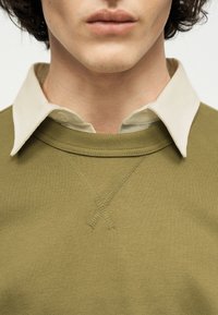 Κοντινό πλάνο ατόμου που φοράει μπλούζα τύπου crewneck σε ελιά πράσινο χρώμα πάνω από μπεζ πουκάμισο με γιακά, δείχνοντας το λαιμό και μέρος του προσώπου.