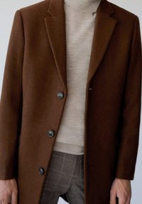 Brown wool coat with a tailored design, notched lapel, and five buttons. Worn over a beige turtleneck and plaid trousers, showcasing textures.