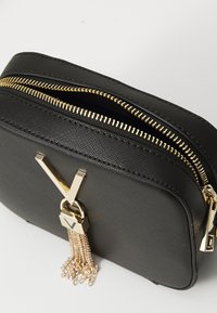 Borsa nera sintetica con doppia cerniera dorata, caratterizzata da un emblema dorato e un dettaglio a nappa. Esterno testurizzato con forma rettangolare.