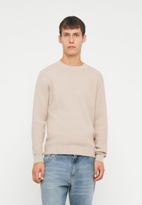 Suéter de punto acanalado beige con mangas largas, cuello redondo y tejido texturizado, combinado con unos vaqueros de denim azul claro.