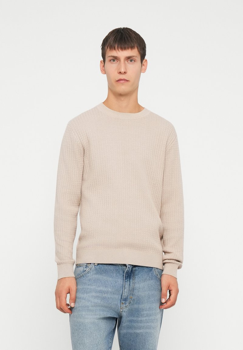 Suéter de punto acanalado beige con mangas largas, cuello redondo y tejido texturizado, combinado con unos vaqueros de denim azul claro.