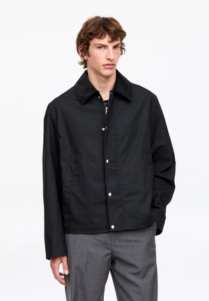 Jeune homme aux cheveux bruns ondulés portant une veste noire boutonnée avec un col en velours côtelé et un pantalon gris, debout sur un fond blanc.