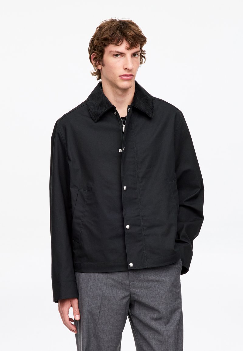 Jeune homme aux cheveux bruns ondulés portant une veste noire boutonnée avec un col en velours côtelé et un pantalon gris, debout sur un fond blanc.