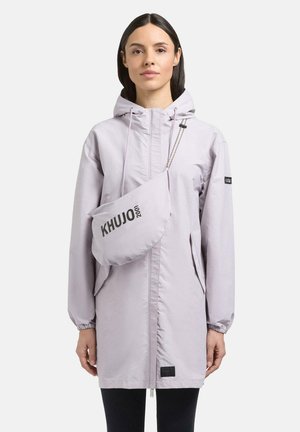 Frau trägt eine hellviolette lange Kapuzenjacke mit passender Umhängetasche mit der Aufschrift „KHUJO 2001“, steht vor einem einfarbigen Hintergrund.