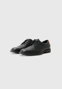 Chaussures de ville en cuir noir avec une texture lisse, bout rond et lacets, présentant une touche de marron à l'arrière du talon.