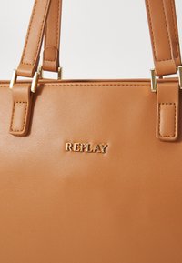 Bruine synthetische handtas met gouden hardware en een "REPLAY" logo. Het ontwerp bevat dubbele handvatten aan de bovenkant en een gladde textuur.