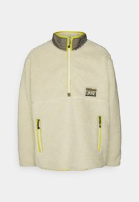 Sudadera de felpa crema con cuello alto, cierre de cremallera y acentos amarillos. Cuenta con dos bolsillos con cremallera y un logo de parche en el pecho.