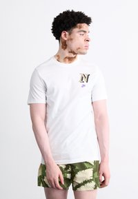 Witte t-shirt met korte mouwen met een gemusterd 'N' logo en een paarse Nike-embleem. Gepaarde met groen en beige gemusterde shorts.