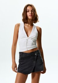 Vit texturerad ärmlös topp med V-ringning och framknutning, kombinerad med en mörkgrå denim minikjol med omslag design och metallknapp.