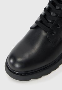 Botte en cuir noir avec un bout arrondi, semelle plate et lacets noirs. Présente une texture lisse et une couture subtile le long du bord.