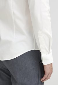 White dress shirt with a curved hem and buttoned cuffs, paired with gray pinstriped trousers, showcasing a smooth texture.