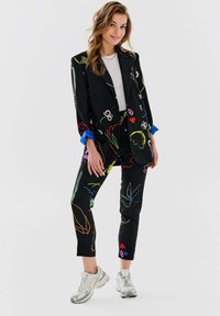 Czarny garnitur dopasowany, z wielokolorowymi wzorami kwiatowymi; zawiera marynarkę z kołnierzykiem i spodnie o wąskim kroju. Materiał wydaje się być fakturowany i mocno ustrukturyzowany.