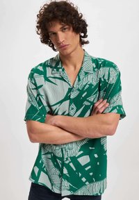 Camicetta verde a maniche corte con un motivo geometrico bianco, realizzata in tessuto leggero, con collo e vestibilità ampia.
