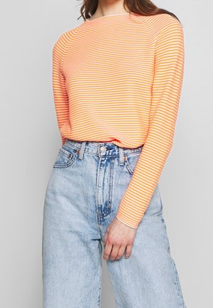 Person wearing long-sleeve orange and white striped top tucked into high-waisted light blue acid-wash jeans, hand resting near hip.