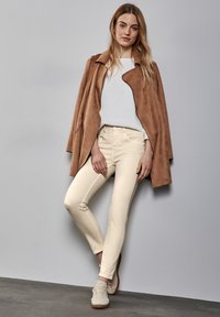 Abrigo de ante marrón, top blanco texturizado, jeans ajustados color beige claro y zapatillas beige con cordones. Diseño simple y colores neutros contra una pared gris.