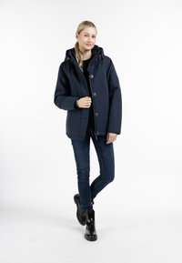 Giacca isolante blu navy con cappuccio, bottoni frontali e due tasche laterali. Un paio di jeans scuri e stivaletti neri alla caviglia completano l'outfit.