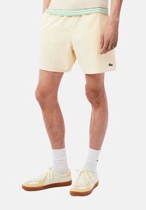 Bañador de cuadros amarillos con una cintura elástica que presenta una franja verde. Se lleva con calcetines blancos y zapatillas crema con acentos marrones.