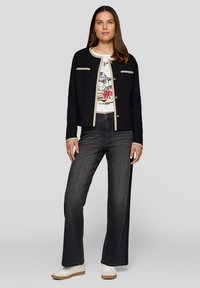 Schwarze, gewebte Strickjacke mit cremefarbenem Saum und Knopfverschluss, kombiniert mit einem Grafik-T-Shirt und dunklen, weiten Jeans, abgerundet mit weißen Turnschuhen.