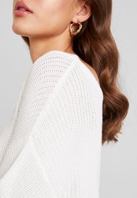 Záběr zblízka na osobu s bílým pleteným svetrem a zkrouceným zlatým kruhovým náušnicím, s vlnitými hnědými vlasy.