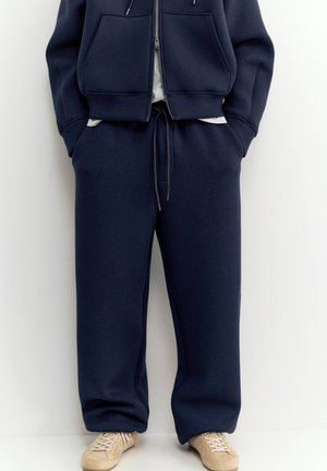 Person trägt dunkelblaue Jogginghose und passenden Kapuzenpullover mit Reißverschluss mit Händen in den Taschen, kombiniert mit beigen Sneakern.