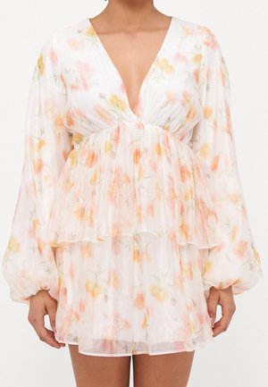 Donna che indossa un vestito bianco trasparente con stampa floreale rosa e gialla, scollo a V profondo, maniche lunghe a sbuffo e gonna a balze.