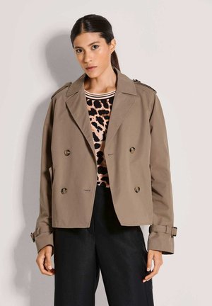 Kaki dubbelgeknoopt trenchcoat met grote knopen, epauletten en opgerolde manchetten, gedragen over een patroon top en zwarte broek.