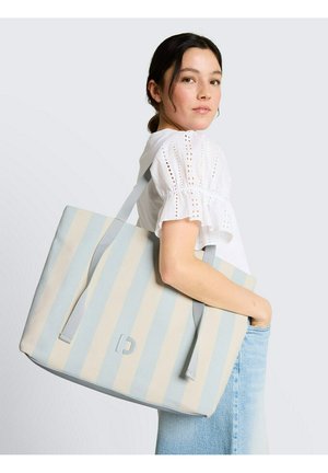 Młoda kobieta w białej bluzce i jasnoniebieskich dżinsach niesie na ramieniu dużą torbę typu tote w niebiesko-beżowe paski.