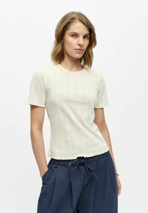 Femme aux cheveux blonds mi-longs portant un haut côtelé crème à manches courtes et un pantalon bleu marine à cordon, debout, une main dans la poche.