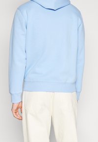 Felpa di cotone azzurro chiaro con tasca a marsupio frontale, polsini e orlo a coste, e cappuccio. Indossata con pantaloni color crema.