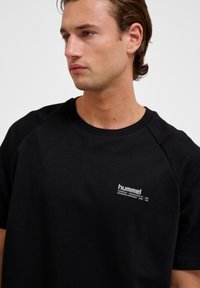 Svart kortärmad t-shirt i bomull med rund halsringning och en liten vit logotyp på övre vänster sida. Slät textur.