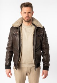 Bruine leren bomberjack met een sherpa kraag, voren rits en meerdere patches. gedragen over een beige trui en lichte broek.