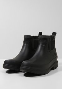 Botas de goma negras hasta el tobillo con paneles laterales elásticos, forro texturizado y suela gruesa. Presenta un logotipo circular en el lado exterior.
