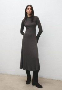 Vestido largo, acanalado y gris oscuro con cuello alto y mangas largas, con una falda acampanada. Combinado con botas altas de ante negro.