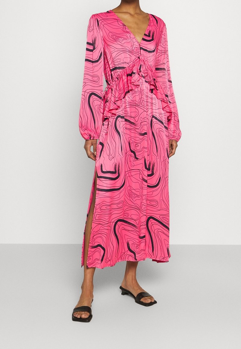 Femme portant une robe à manches longues rose vif avec des motifs tourbillonnants abstraits noirs et des volants à la taille, associée à des sandales noires à bout ouvert.