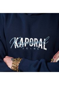 Sudadera azul marino con el logotipo blanco de "Kaporal Jeans"; el tejido parece suave. Se ven manos con pulseras de cadena doradas en la parte inferior.
