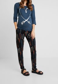 Modrá košile s dlouhým rukávem a designem metalické srdce a šipky, doplněná černými kalhotami s barevnými vertikálními pruhy a černými sandály.