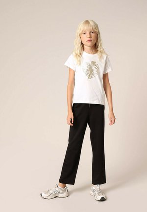 T-shirt en coton blanc avec un motif de cœur doré métallique et d'éclair, associé à un pantalon noir ajusté et des baskets grises.