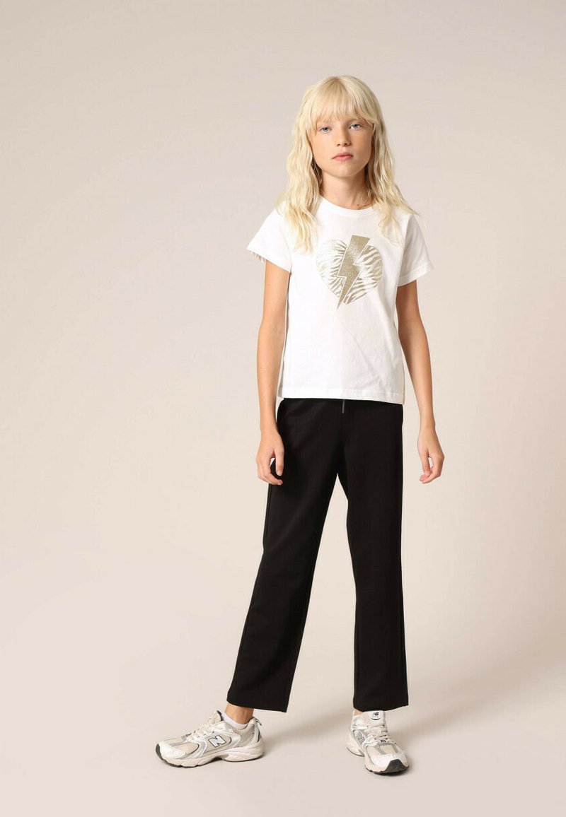 T-shirt en coton blanc avec un motif de cœur doré métallique et d'éclair, associé à un pantalon noir ajusté et des baskets grises.