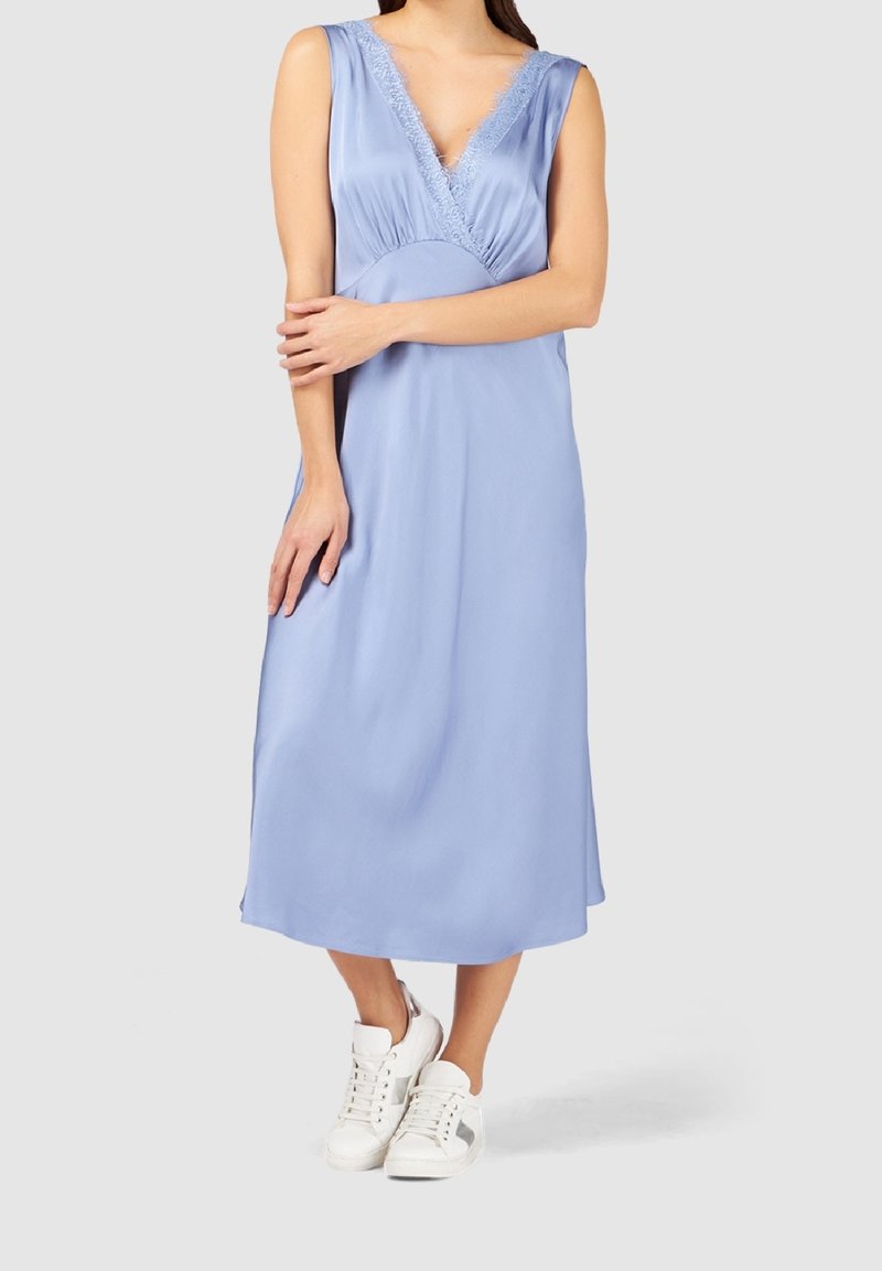 Femme portant une robe midi bleu clair sans manches avec un bord en dentelle et des baskets blanches, debout les bras croisés sur le torse.