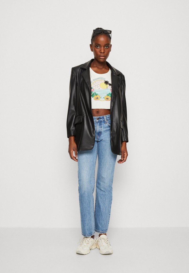 Schwarze, oversized Blazer aus Kunstleder über einem weißen, kurzen Top mit buntem, tropischem Druck, kombiniert mit hellblauen, geraden Jeans und Sneakers.