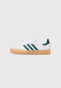 White sneakers with green stripes, gum sole, and lace-up design. Smooth upper material with a textured rubber sole. Classic silhouette.