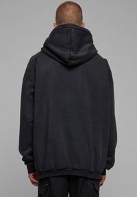 Persona con capelli corti che indossa un ampio hoodie nero e pantaloni cargo neri, voltata di spalle contro uno sfondo grigio semplice.