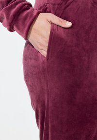 Měkká, burgundská látková oděv s boční kapsou. Textura vypadá jemně a hladce. Design zahrnuje dlouhé rukávy.