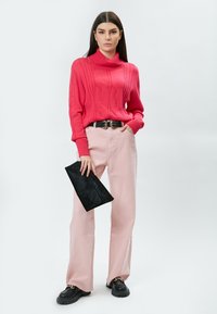 Pull en tricot rose à col montant avec un motif de tresses, pantalon rose clair taille haute, sac à main noir, et chaussures noires épaisses avec une semelle épaisse.